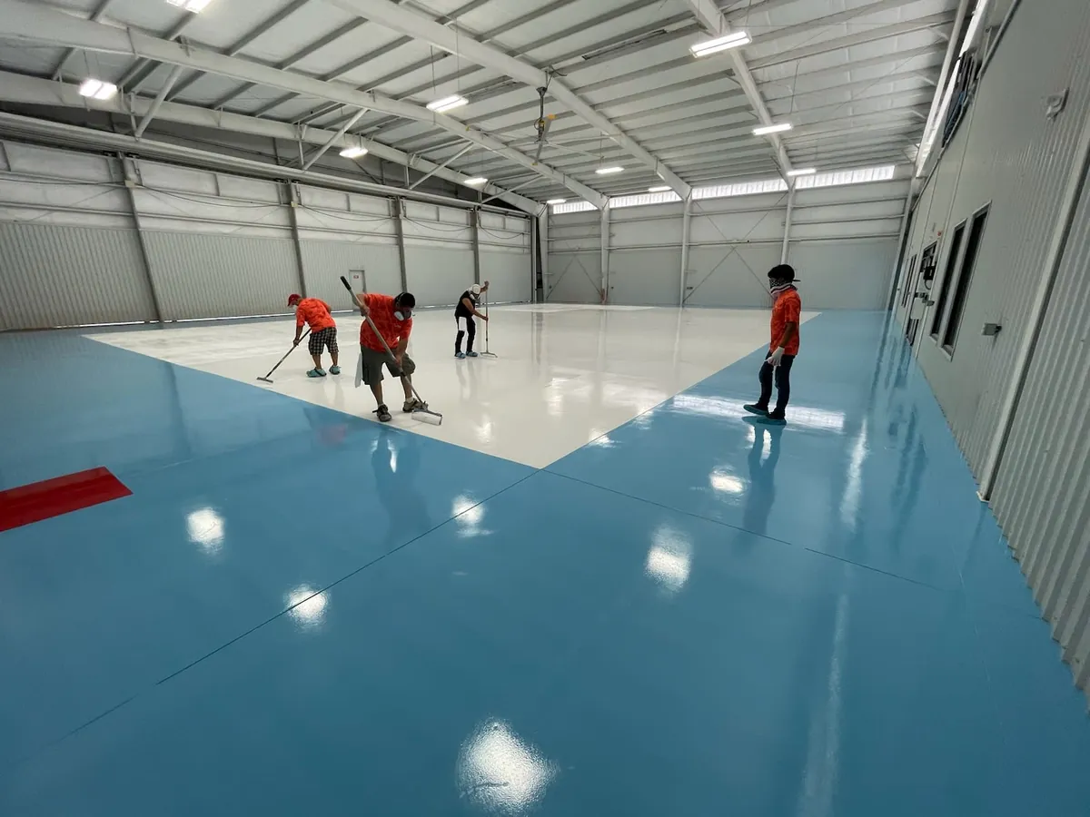 Crew rolling a white coat across a blue base coat on a large aircraft hangar floor