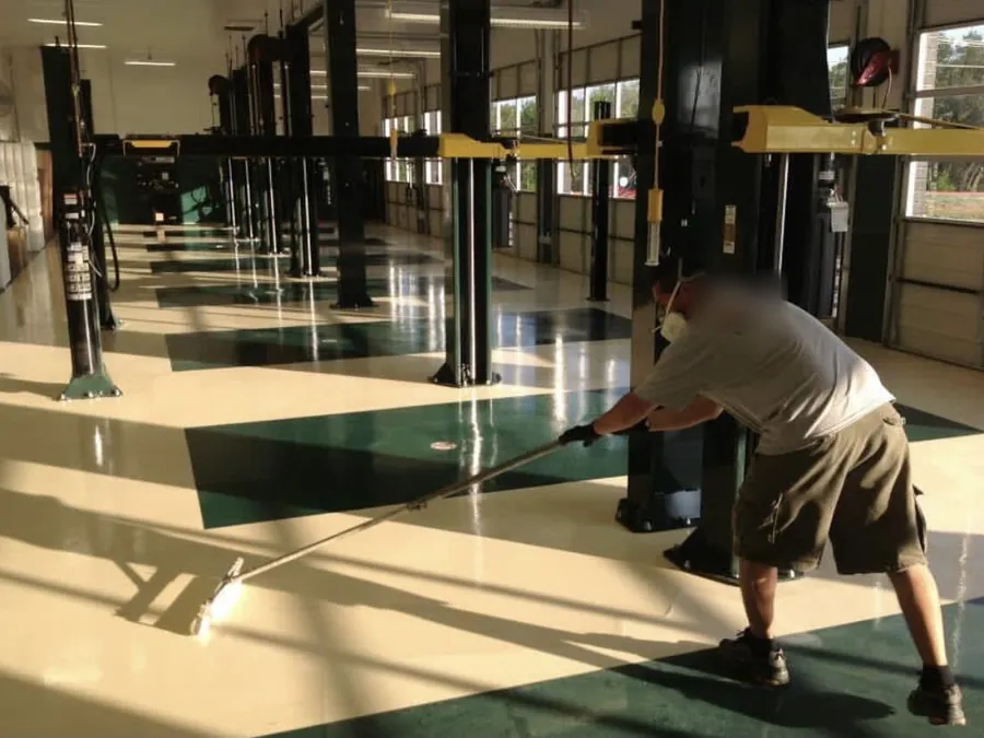 Crew member rolling a clear topcoat over a cream and green auto shop floor with lifts